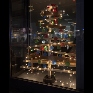 Ein aus Brettern gezimmerter Weihnachtsbaum, der in einem dunklen Schaufenster steht und mit einer leuchtenden Lichterkette geschmückt ist. An dem "Baum" hängen Weihnachtsbaumkugel aus Papier, auf denen handschriftliche Dinge stehen.