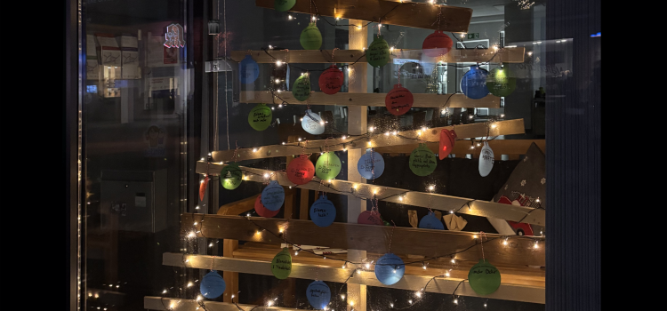 Ein aus Brettern gezimmerter Weihnachtsbaum, der in einem dunklen Schaufenster steht und mit einer leuchtenden Lichterkette geschmückt ist. An dem "Baum" hängen Weihnachtsbaumkugel aus Papier, auf denen handschriftliche Dinge stehen.